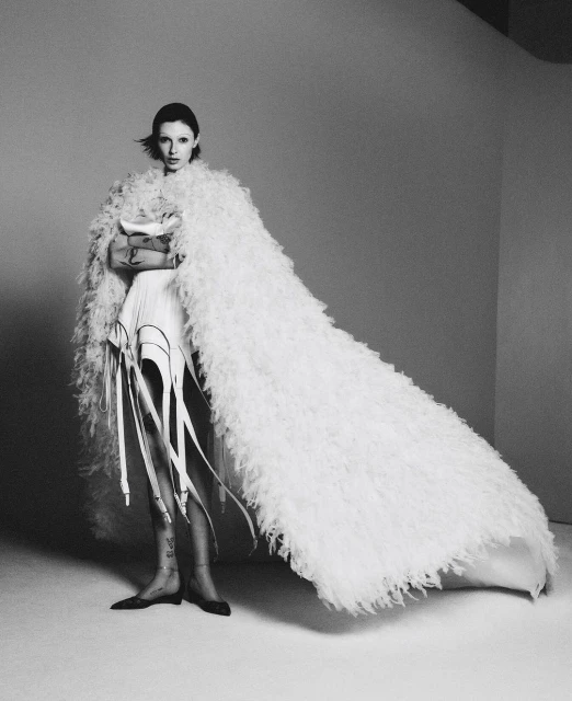 Black and white portrait of a fashionable adult woman in an off-white shaggy cloak, accessorized with leg garters in a studio setting.