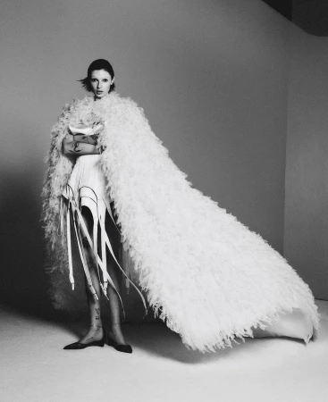 Black and white portrait of a fashionable adult woman in an off-white shaggy cloak, accessorized with leg garters in a studio setting.