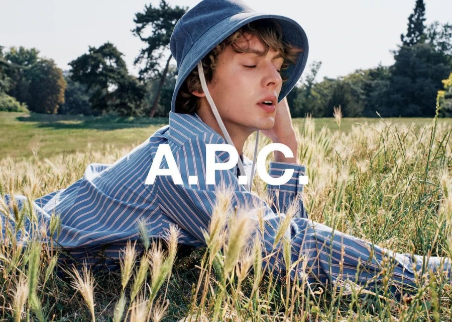A blond male fashion model poses in a field wearing a blue and white striped shirt and matching hat, with the APC logo visible.