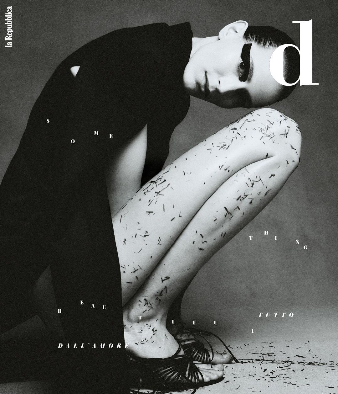 Monochrome fashion shot of a woman crouching with dark hair, a black jacket, and dark shoes, scattered leaf debris, and magazine text.