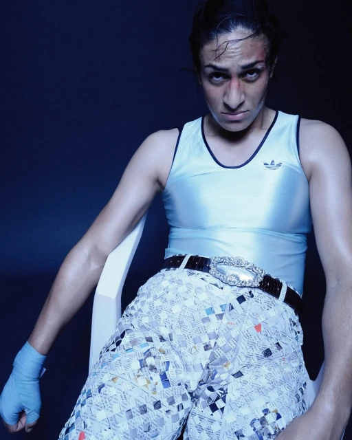 Boxer in avant-garde fashion, seated in a blue-lit studio. Athletic woman wears a shiny tank top, sequined shorts, and boxing wraps.