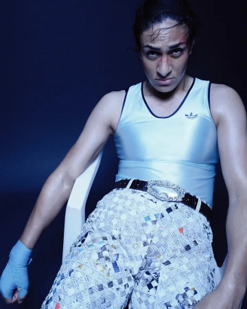 Boxer in avant-garde fashion, seated in a blue-lit studio. Athletic woman wears a shiny tank top, sequined shorts, and boxing wraps.