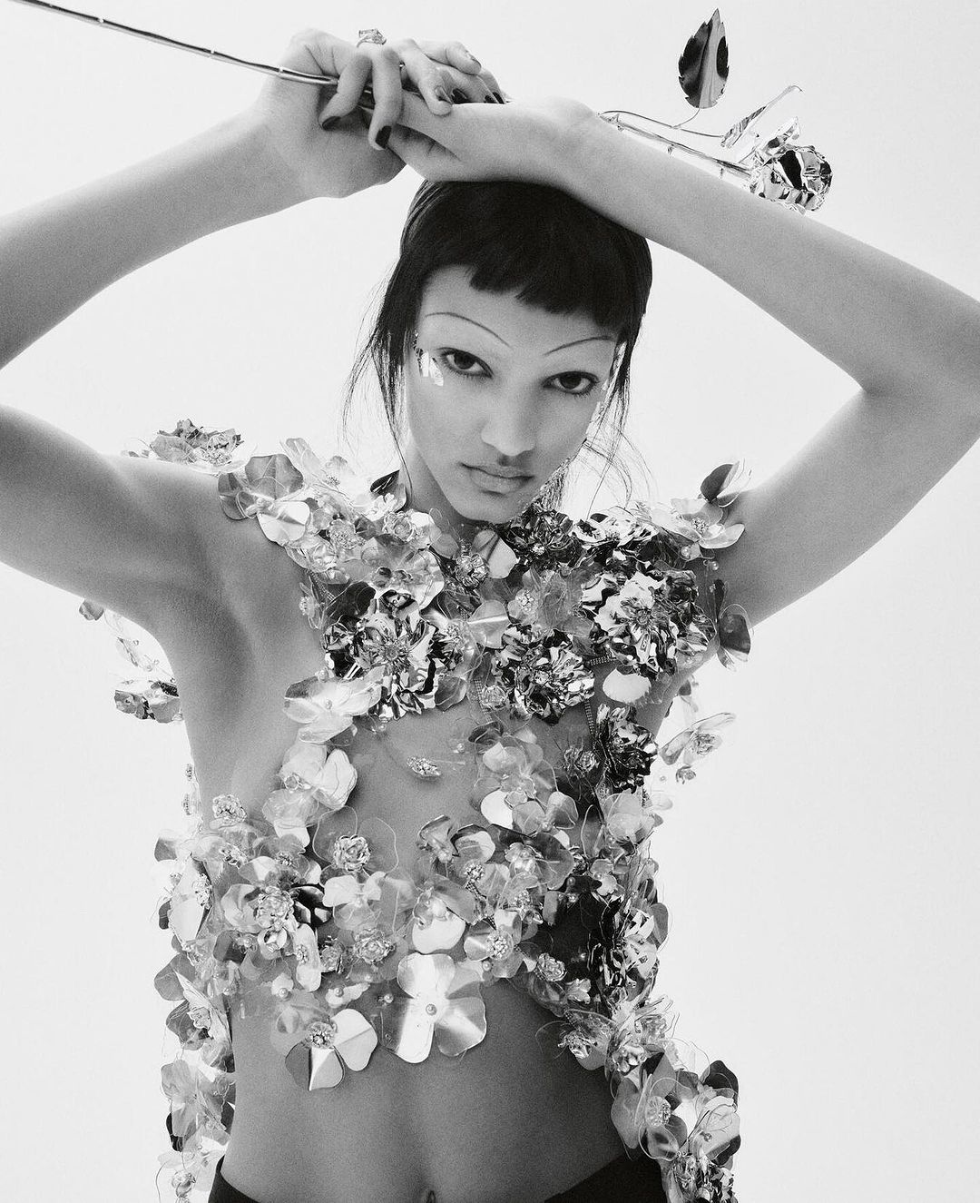 Black and white fashion portrait of a woman with short dark hair, wearing silver floral embellishments, holding a metallic rose.
