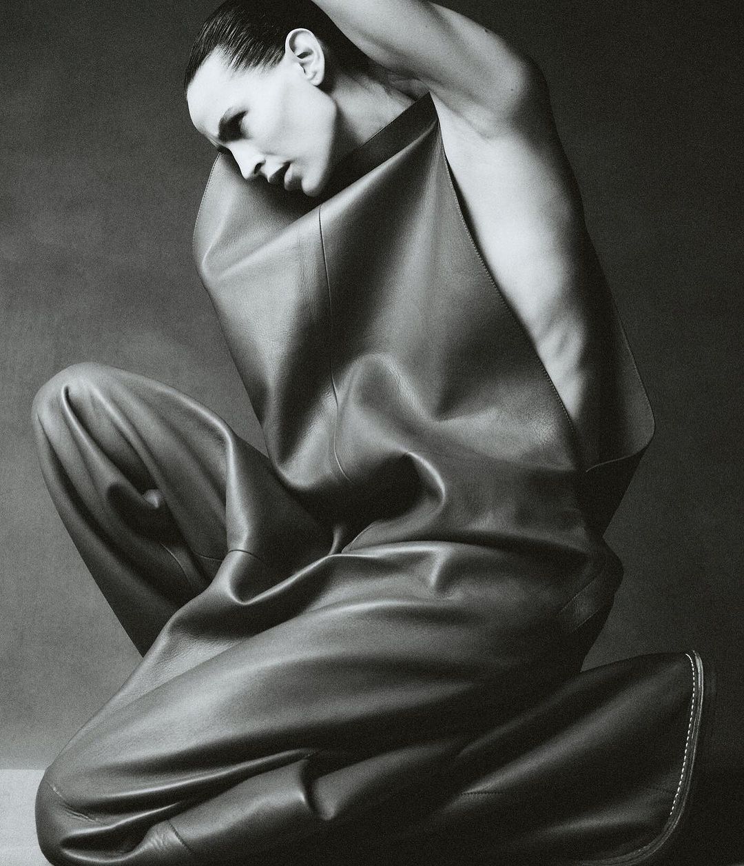 Monochrome studio portrait of a fashion model in leather clothing, kneeling with hands behind head against a grey backdrop.