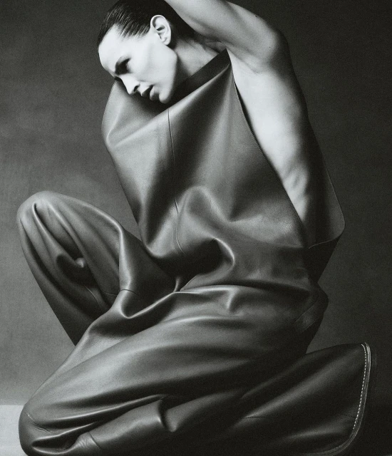 Monochrome studio portrait of a fashion model in leather clothing, kneeling with hands behind head against a grey backdrop.