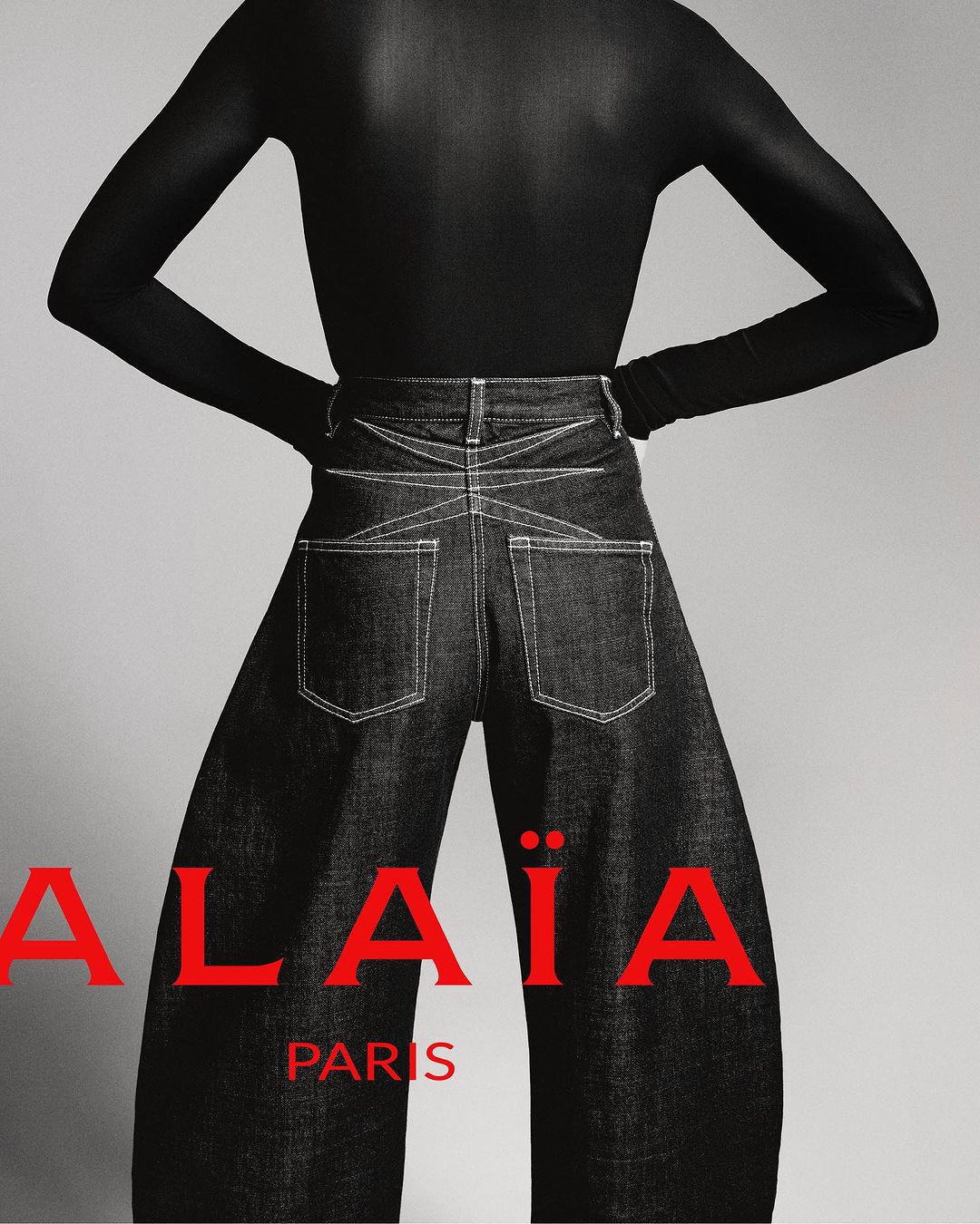 Fashion shot featuring a model in a black top and wide-leg denim pants, posing against a neutral background. The brand ALAÏA is visible.
