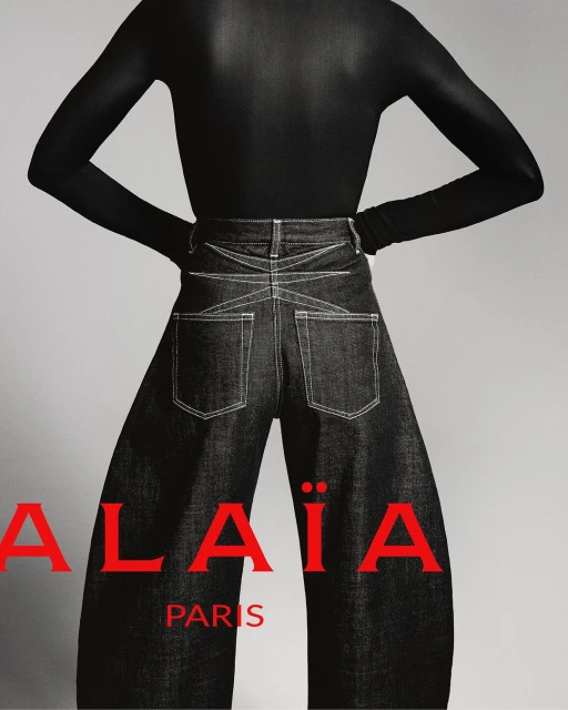 Fashion shot featuring a model in a black top and wide-leg denim pants, posing against a neutral background. The brand ALAÏA is visible.
