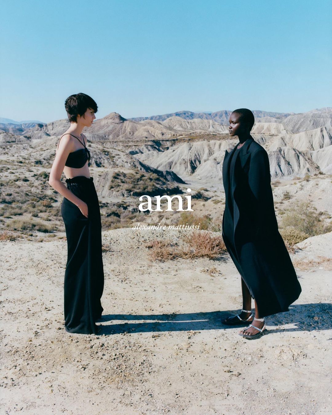 Two fashion models pose outdoors in black attire against a desert backdrop under a clear sky, showcasing minimalist and elegant style.