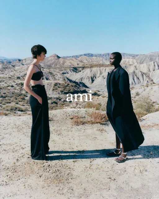 Two fashion models pose outdoors in black attire against a desert backdrop under a clear sky, showcasing minimalist and elegant style.