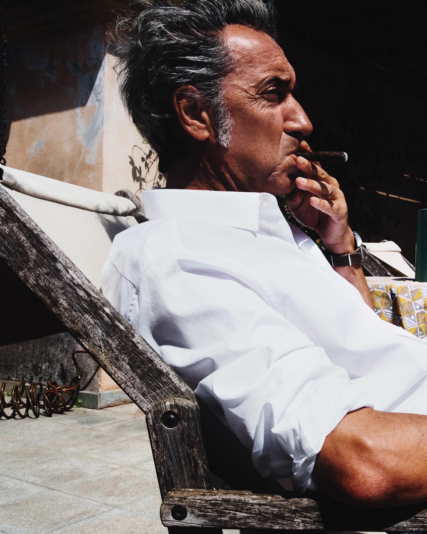 Profile shot of an adult male in a white shirt, smoking a cigar while sitting in a wooden chair outdoors with sunlight and shadows.