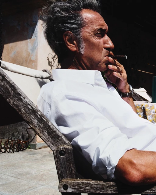 Profile shot of an adult male in a white shirt, smoking a cigar while sitting in a wooden chair outdoors with sunlight and shadows.