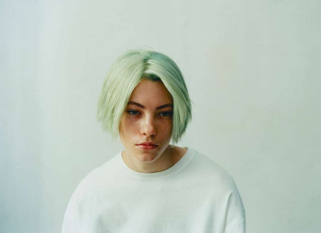 Close-up portrait of a young adult female with short, light-green hair, wearing a white sweatshirt, against a pale-colored wall indoors.