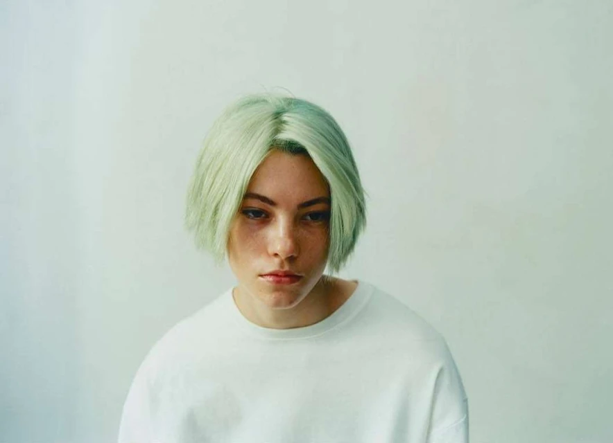 Close-up portrait of a young adult female with short, light-green hair, wearing a white sweatshirt, against a pale-colored wall indoors.