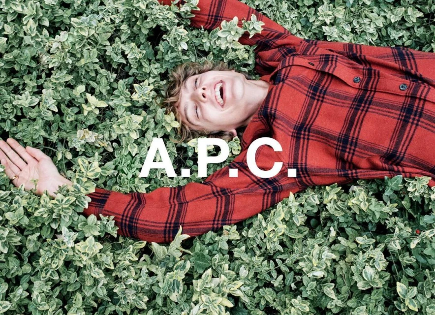 Adult male lying on a bed of green plants. He is wearing a red and black plaid shirt. The A.P.C. logo is superimposed.