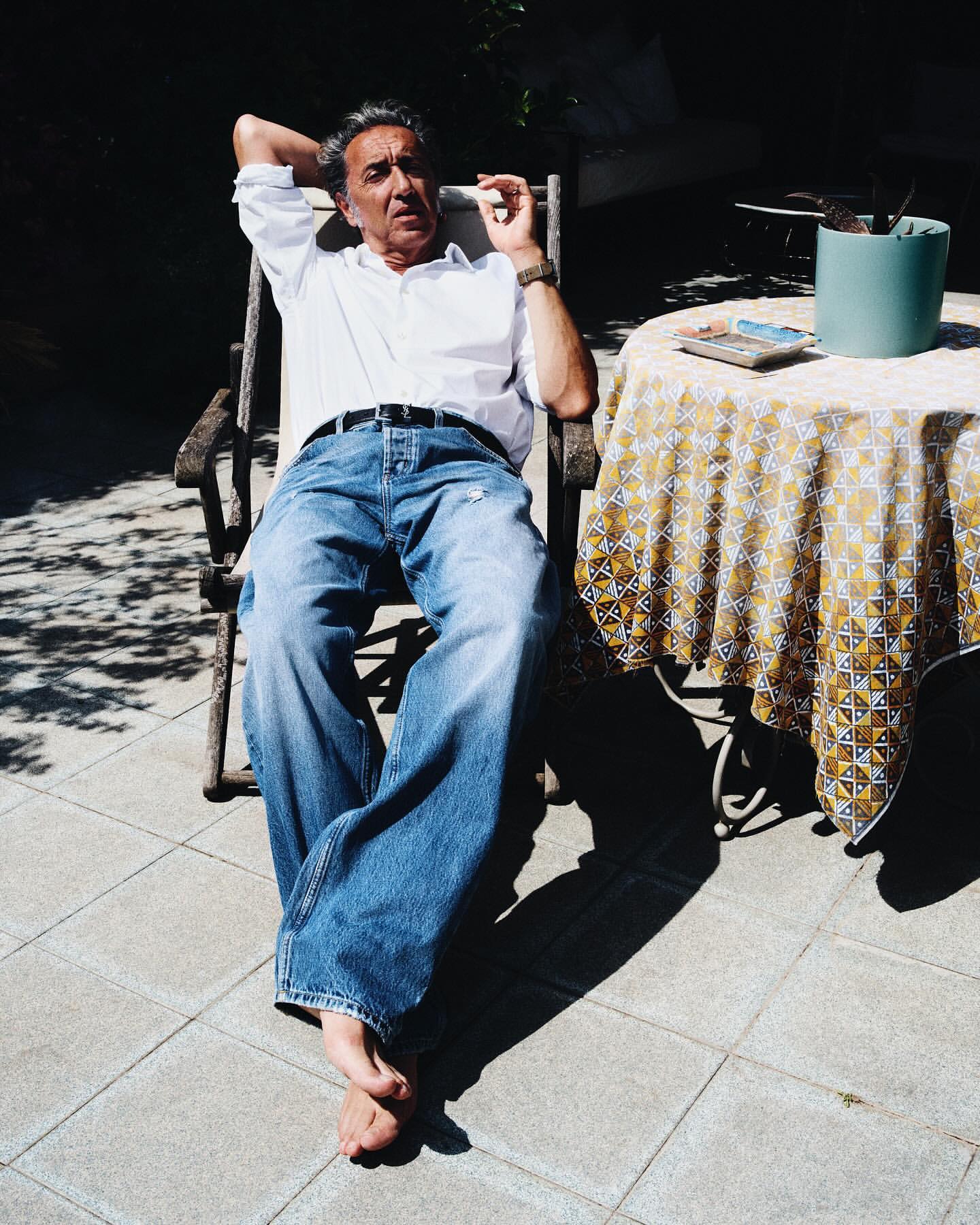 Adult man relaxing outdoors in sunlight. He is sitting in a wooden chair on a tiled patio with a table nearby, with a cigarette.