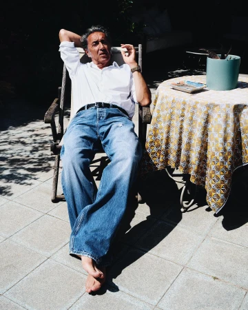 Adult man relaxing outdoors in sunlight. He is sitting in a wooden chair on a tiled patio with a table nearby, with a cigarette.