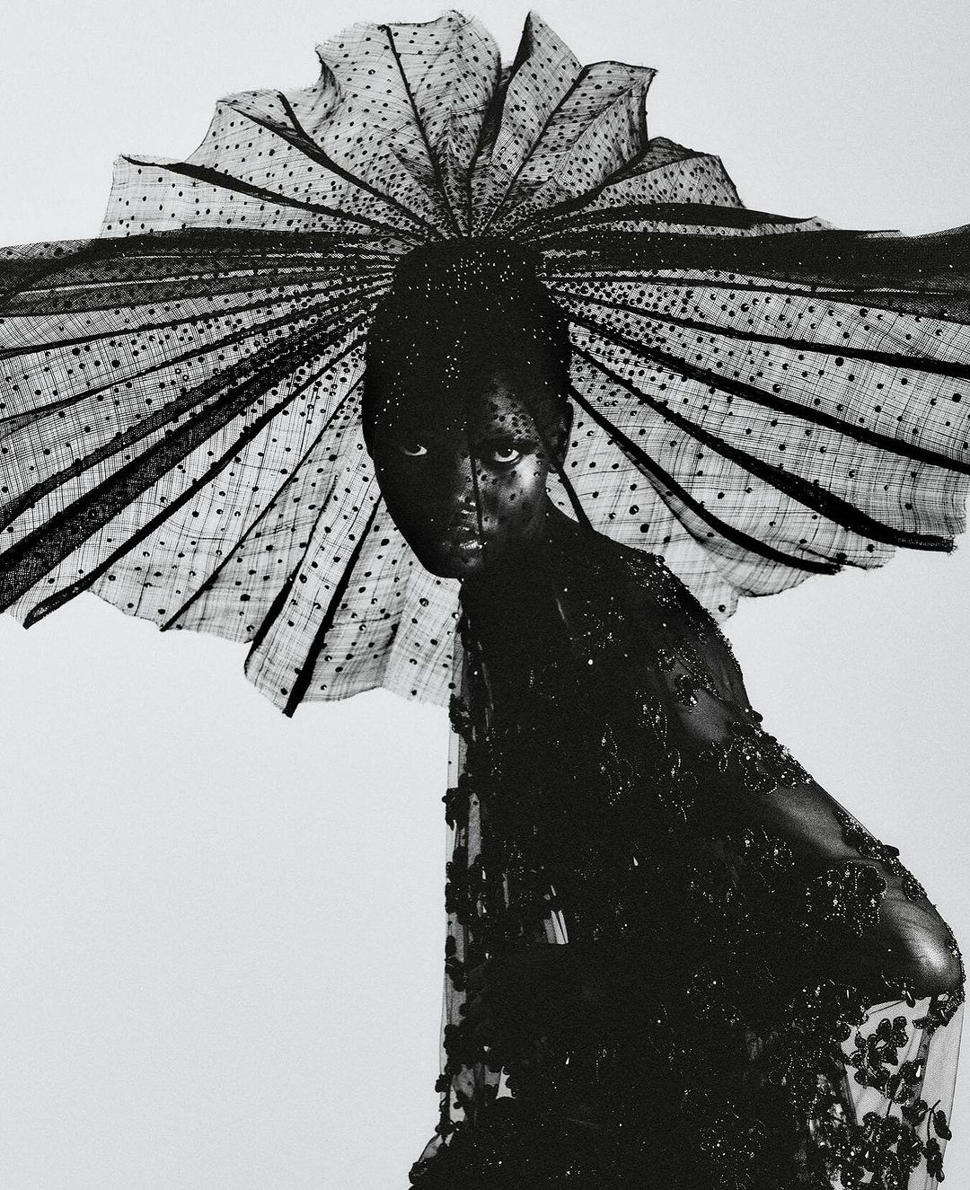 Woman in an elaborate black beaded and lace dress with matching headpiece posing against a white background in a fashion shot.