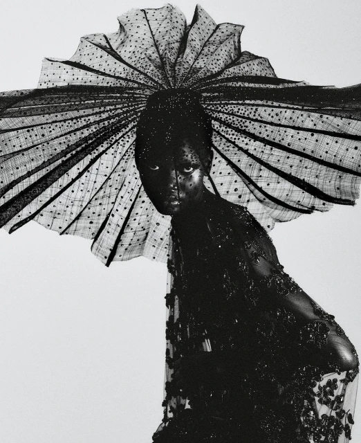 Woman in an elaborate black beaded and lace dress with matching headpiece posing against a white background in a fashion shot.