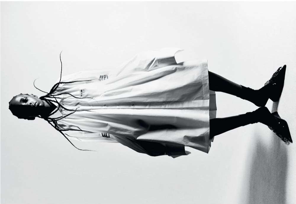 Fashion editorial shot of a Black woman in a white coat and black high heels, lying on a reflective surface against a white backdrop.