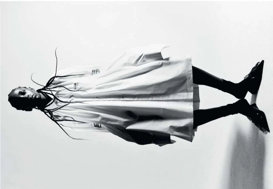 Fashion editorial shot of a Black woman in a white coat and black high heels, lying on a reflective surface against a white backdrop.