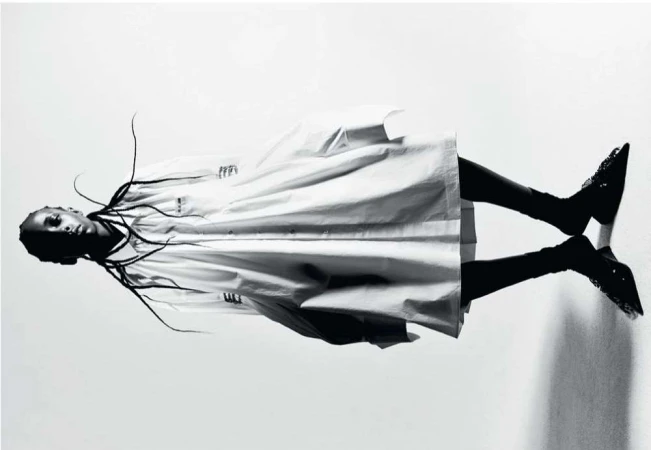 Fashion editorial shot of a Black woman in a white coat and black high heels, lying on a reflective surface against a white backdrop.