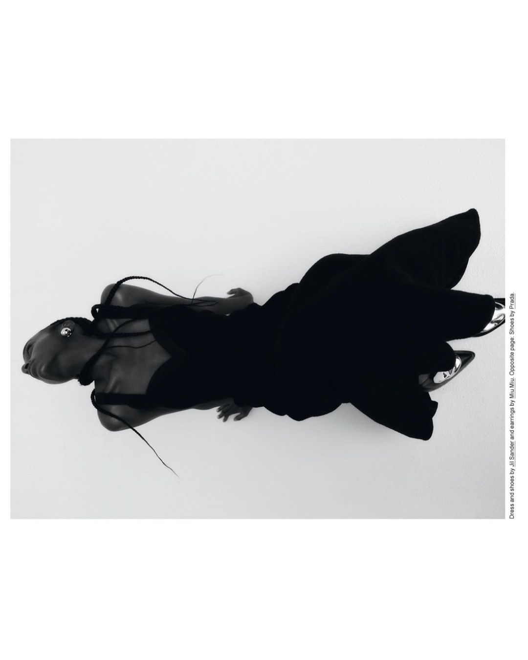Top-down editorial fashion shot: Woman in a black gown and silver heels lies on a white surface with soft studio lighting.  