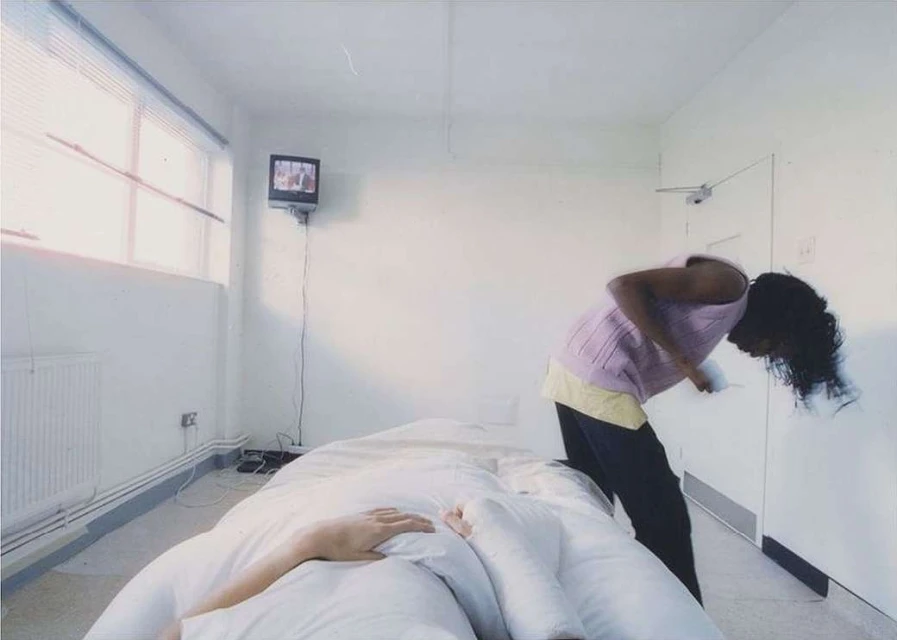 Indoor scene featuring an adult female bending over a bed with a person lying underneath the white duvet, and a television in the corner.
