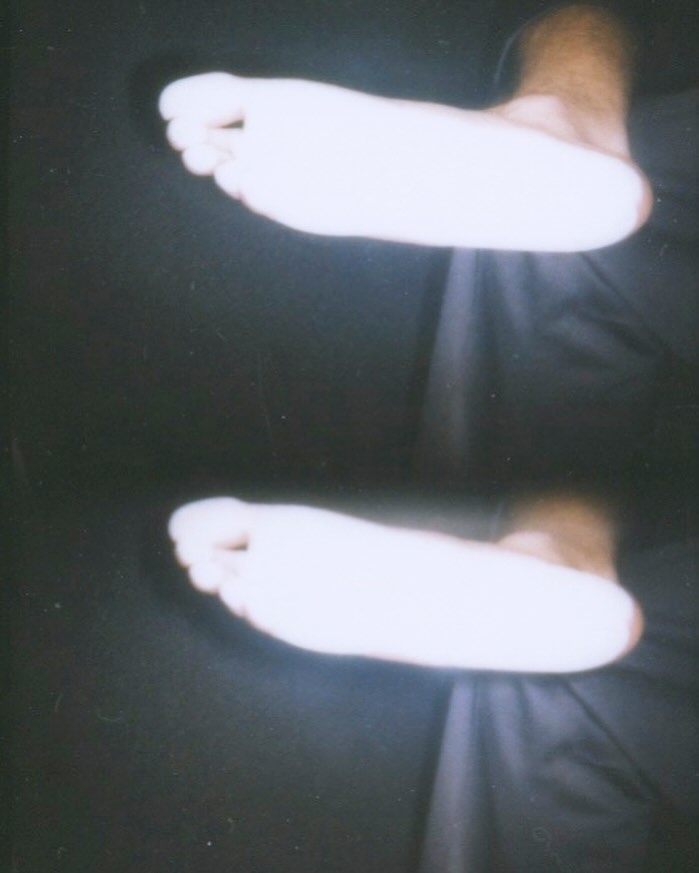 Low-angle shot of two translucent human feet in a stacked vertical composition against a dark, shadowy background. Minimalist aesthetic.