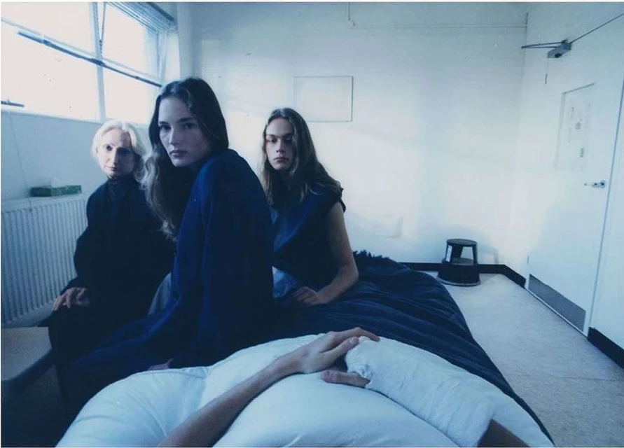 Three women in blue dresses are pictured indoors, one elderly woman and two young adult women with a white bed in the foreground.