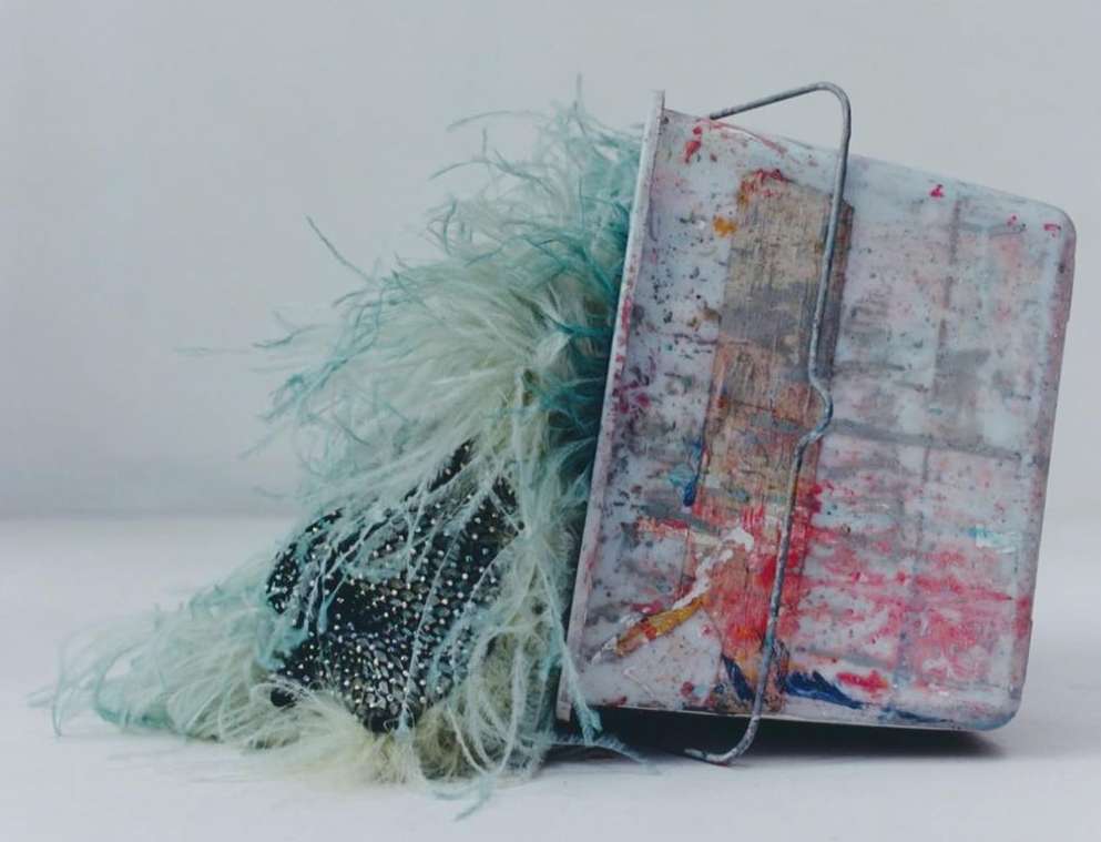 Still life image featuring a container with a metal handle, adorned with blue feathers and a beaded textile, set against a plain backdrop.