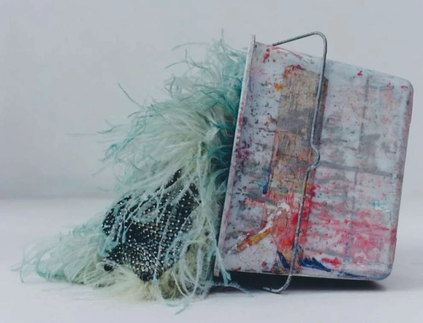 Still life image featuring a container with a metal handle, adorned with blue feathers and a beaded textile, set against a plain backdrop.