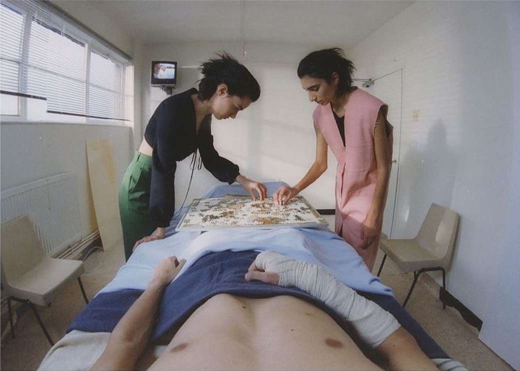 Two women completing a puzzle for a man lying on an examination table in a clinical room. Patient is partially covered with a blanket.