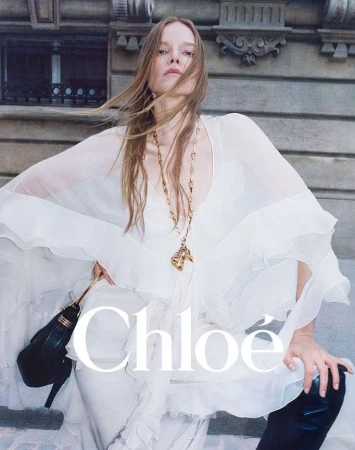 Model in a flowing white blouse holding a black handbag against an exterior stone building backdrop