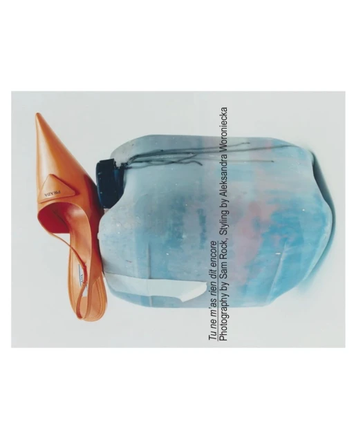 Still life of an orange Prada heel leaning against a light blue sculpture on a white background, minimalist fashion photography.