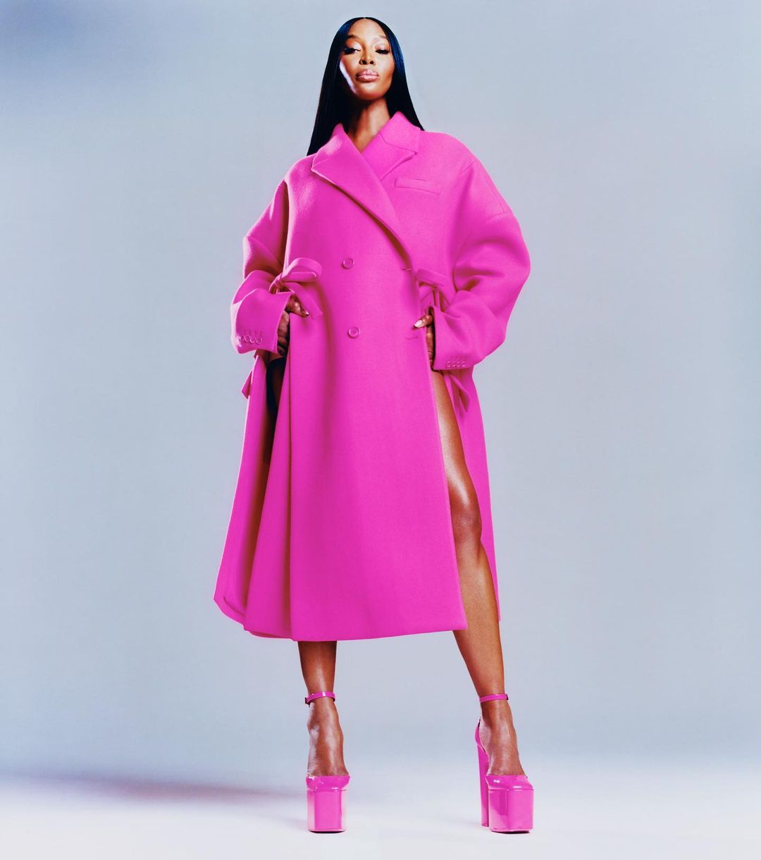 Naomi Campbell poses in a vibrant pink double-breasted overcoat and matching platform heels against a pale blue background in a studio shot.