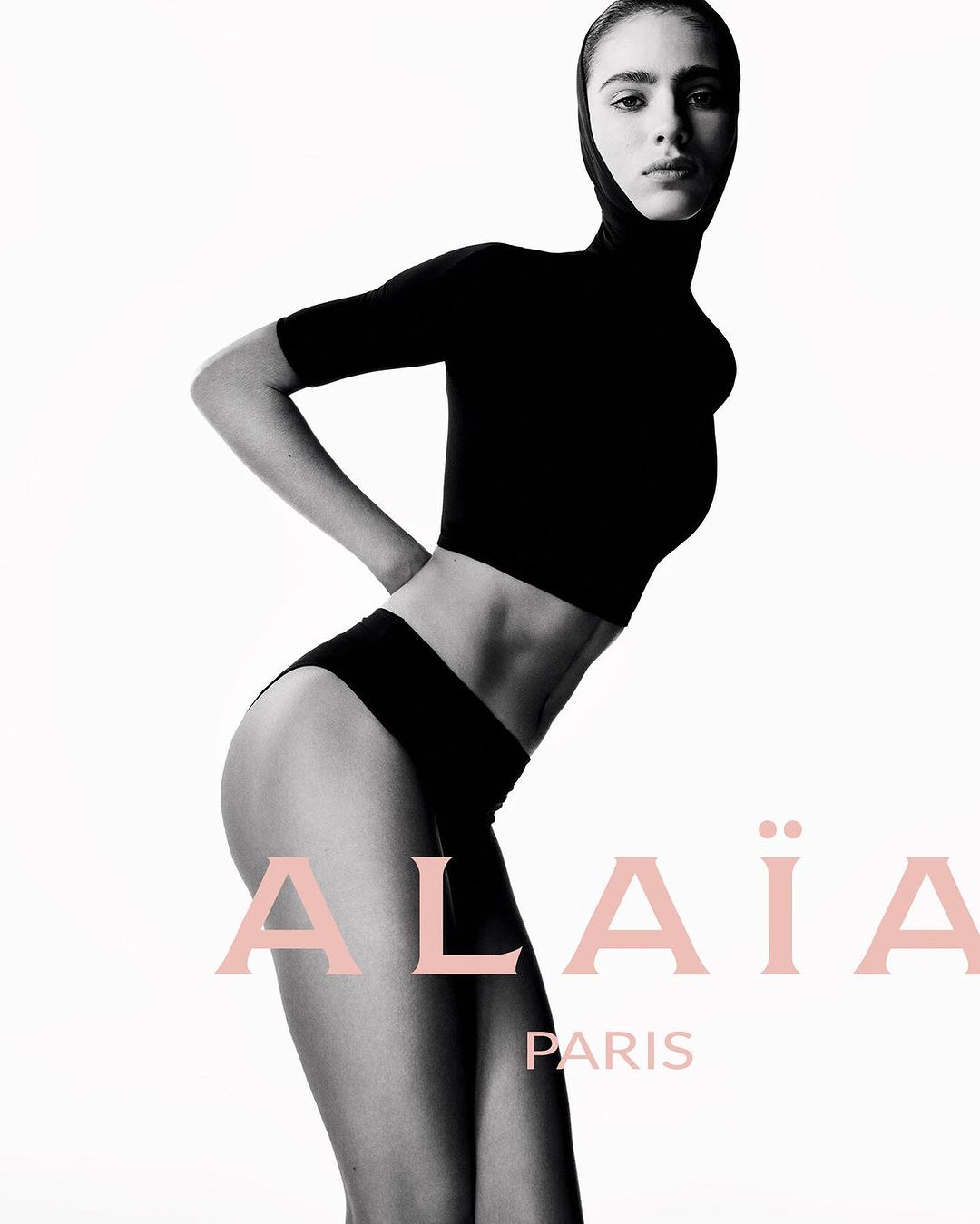 Fashion shot of a model wearing a black top and briefs against a white backdrop. The Alaïa Paris logo is visible.