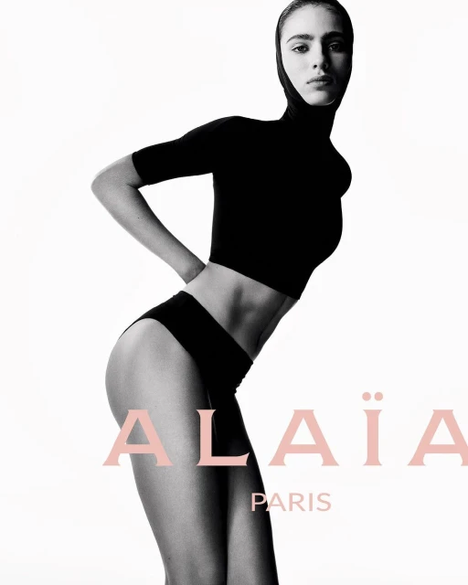 Fashion shot of a model wearing a black top and briefs against a white backdrop. The Alaïa Paris logo is visible.