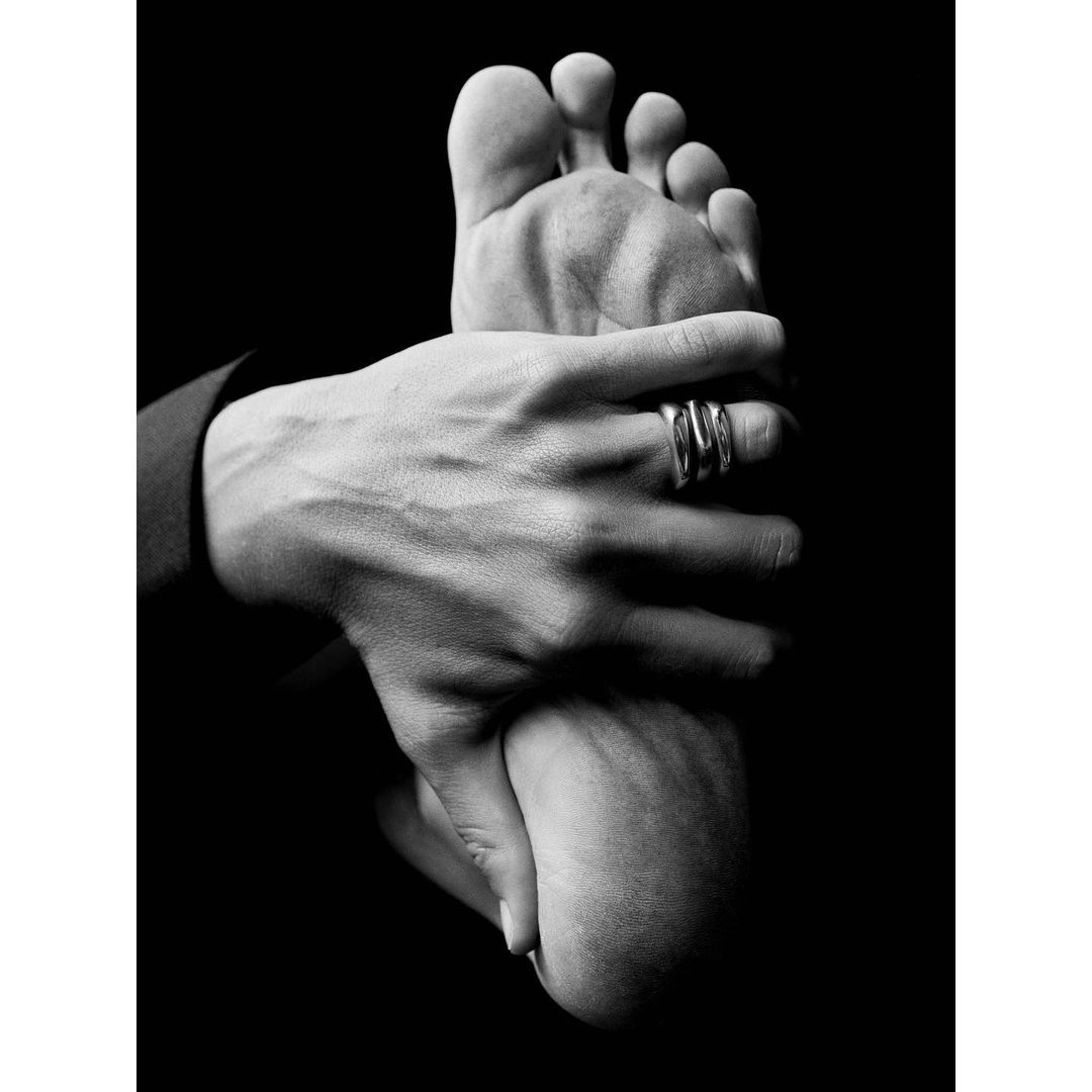 Monochrome photo of a person's hand holding a foot, showcasing a silver ring on the hand against a stark black backdrop in a studio setting.