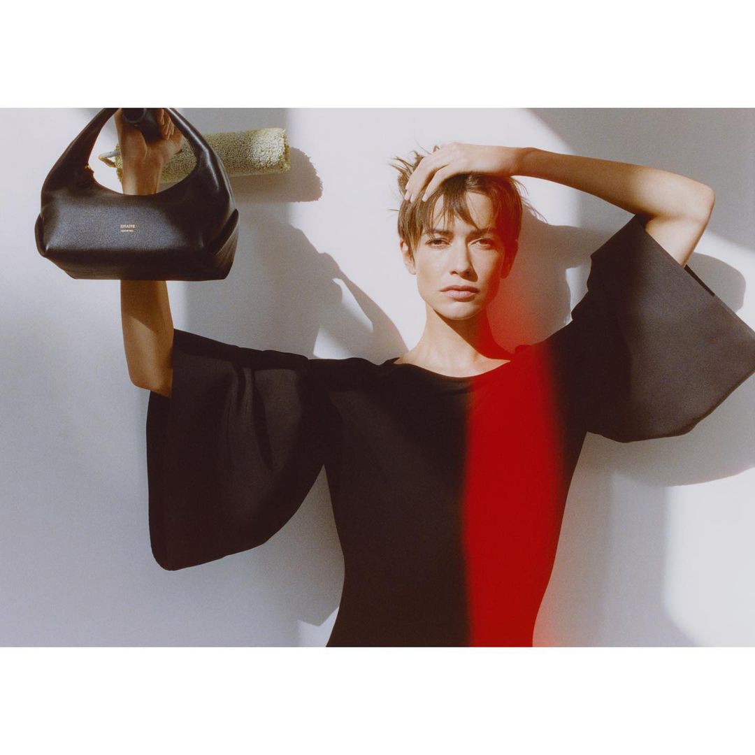 Fashion shot of a woman holding a black handbag in a bright, naturally lit studio with a red light effect over her torso.