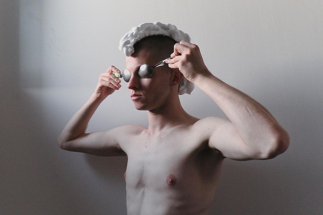 Conceptual portrait of shirtless man with shaving foam on his head, spoons covering his eyes, and holding a razor inside an indoor setting.