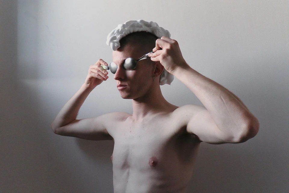 Conceptual portrait of shirtless man with shaving foam on his head, spoons covering his eyes, and holding a razor inside an indoor setting.