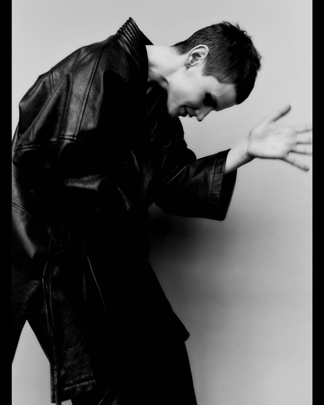Monochromatic portrait of a young adult male in a leather jacket, bending forward with an open hand against a light grey backdrop.