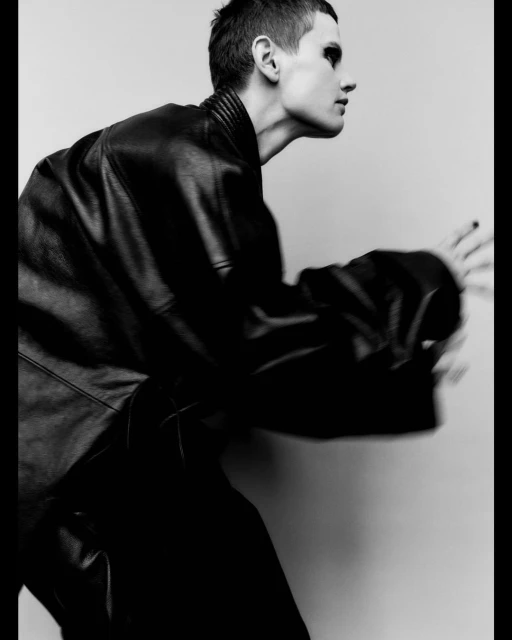 Monochrome studio shot of an androgynous adult in motion, wearing a leather jacket, with dark eye makeup and short hair, against a grey background.