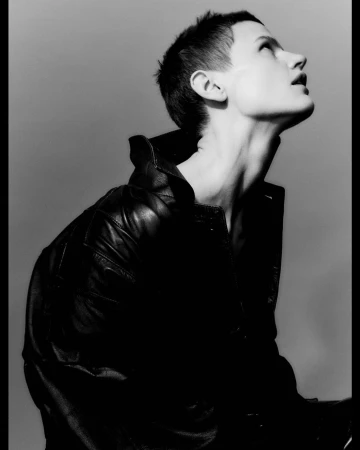 Monochrome portrait of a short-haired woman in a black leather jacket looking upward against a grey gradient background.