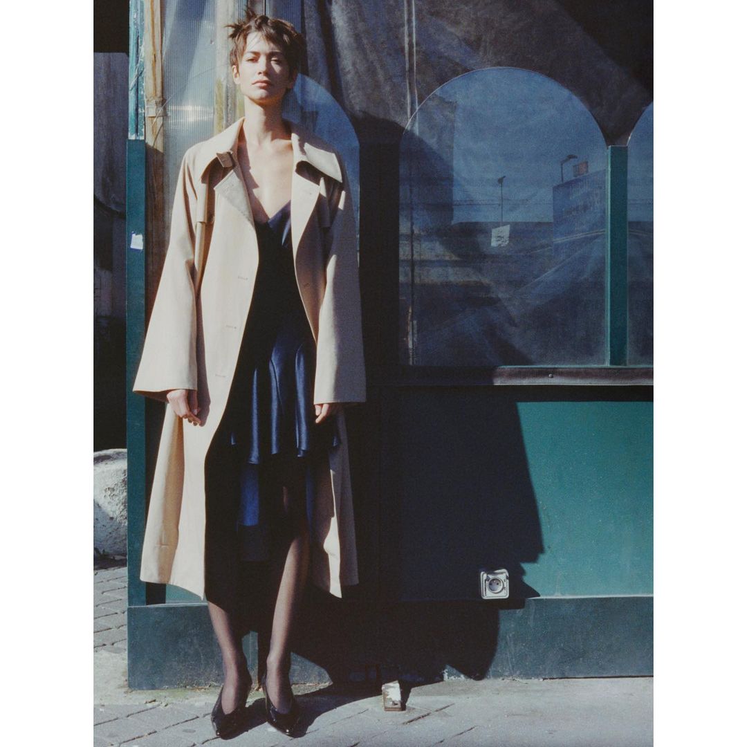 Model standing outdoors wearing an open beige trench coat over a dark dress and dark stockings against a green wall