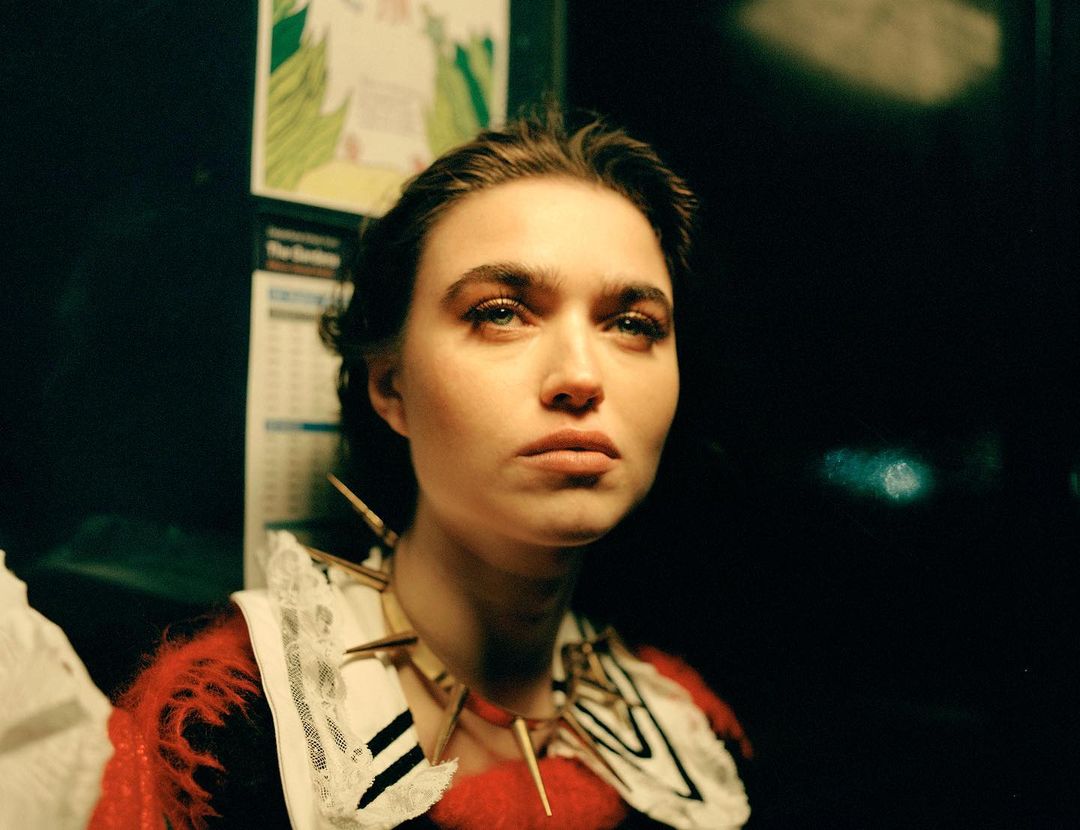 Portrait of a woman with fair skin and dark hair, wearing a white and red garment with decorative neck piece, against a dark background.