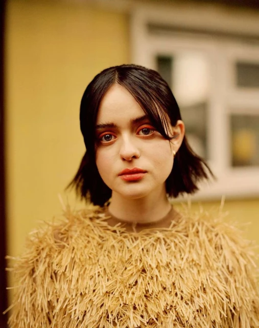 Portrait of a young woman with short dark hair, wearing a textured gold sweater, set against a pale yellow wall with a window.