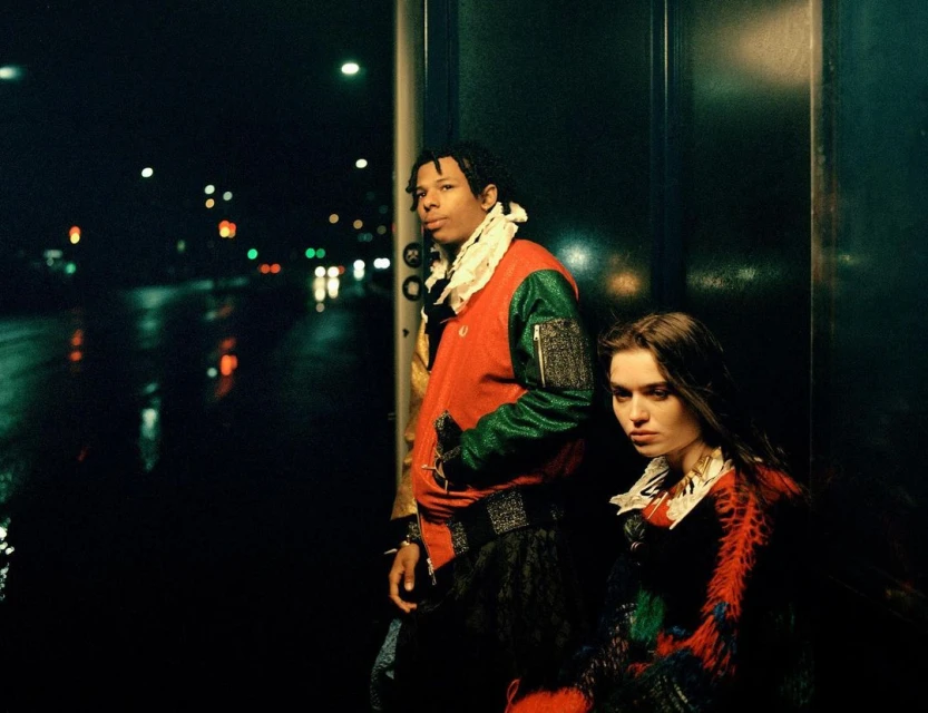 Two people in colorful jackets stand at night against a dark backdrop with blurred city lights, reflecting in the wet ground.