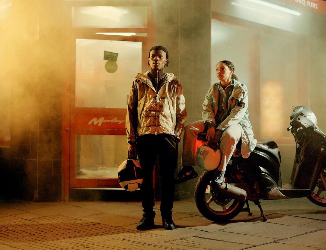 Two people pose in urban setting, motorcycle parked nearby. Man stands holding helmet, woman sits on motorcycle, wearing light-colored jackets.