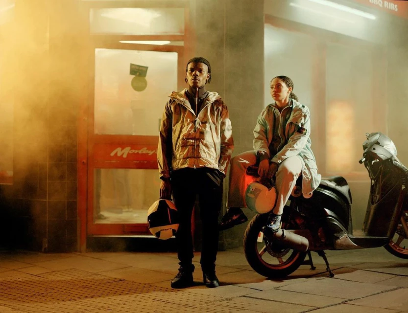 Two people pose in urban setting, motorcycle parked nearby. Man stands holding helmet, woman sits on motorcycle, wearing light-colored jackets.